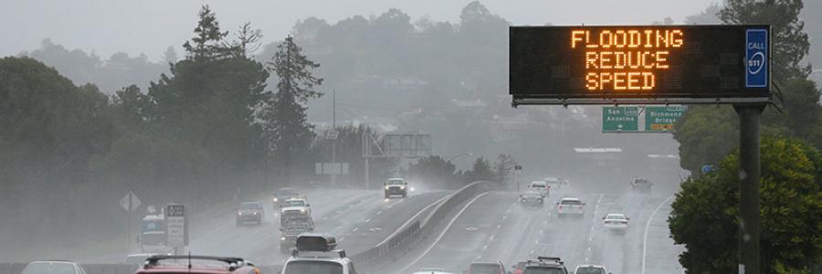 Cars driving on the freeway in rain with an electric sign saying "Flooding Reduce Speed"