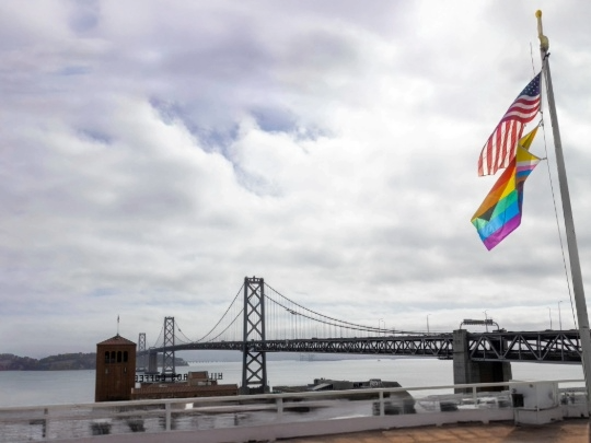A view of the Bay Bridge with the American and Pride flags waving in the foreground