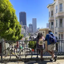 Two people next to a bicycle peer down over a railing at a bustling cityscape below