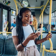 A smiling woman wearing headphones grips a bus handrail and smiles at her smartphone as she rides the bus