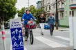 A mother pedals a bicycle with two children riding behind her, while two pedestrians walk behind, all in front of an SFMTA Slow Streets sign