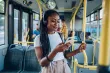 A smiling woman wearing headphones grips a bus handrail and smiles at her smartphone as she rides the bus