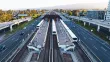 BART trains at a center platform station between freeway lanes in the San Francisco Bay Area, with cars on either side and hills in the background at sunset.